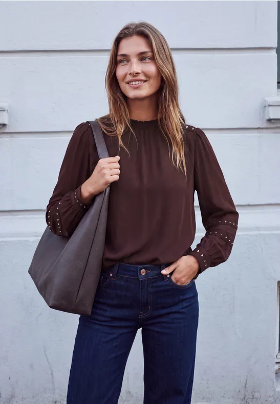 Une femme souriante porte un chemisier marron, un jean bleu et un sac gris devant un mur clair.