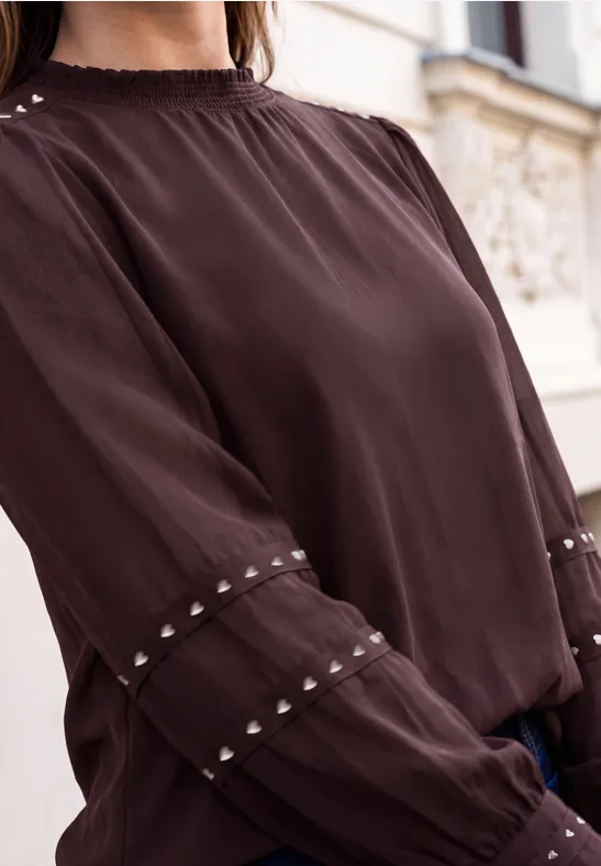 Blouse brun foncé à manches longues avec ornements clous aux poignets, portée devant un bâtiment.
