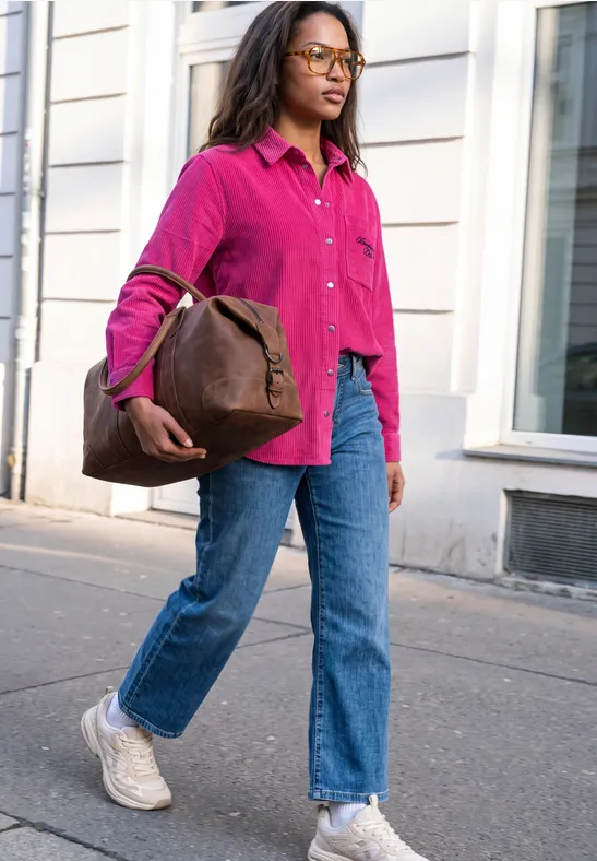 Frau in pinker Jacke, Jeans und weißen Sneakers trägt braune Tasche und geht auf Gehweg.