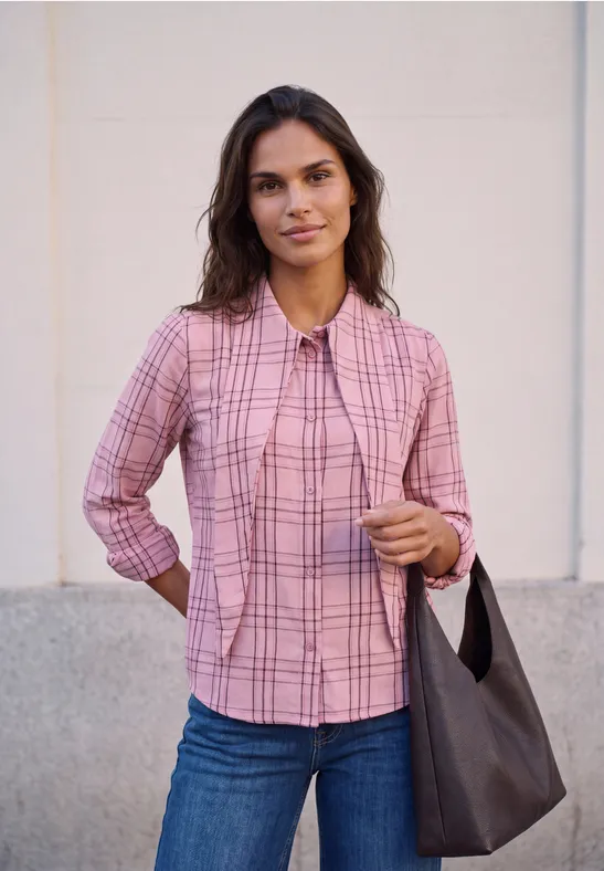 Frau in rosa kariertem Hemd und Jeans mit dunkelgrauer Tasche lächelt in die Kamera.