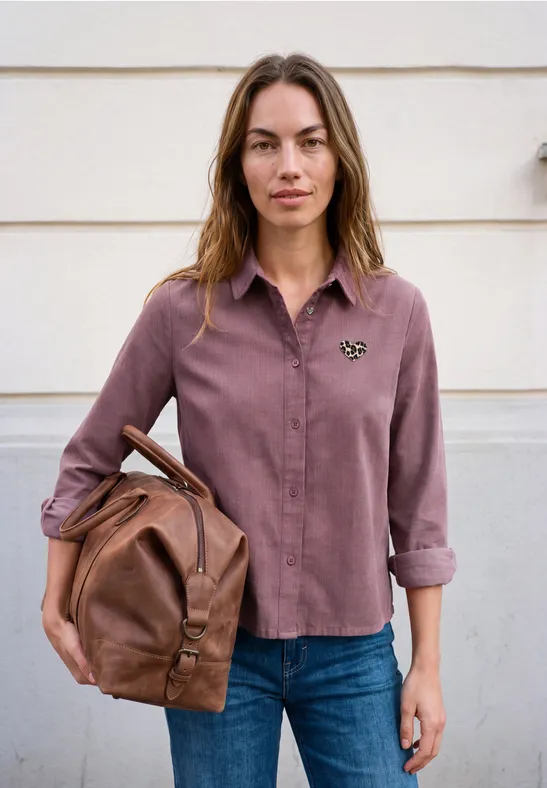 Eine junge Frau steht vor einer hellen Wand, trägt blaue Jeans, eine violette Bluse und hält einen braunen Lederrucksack.