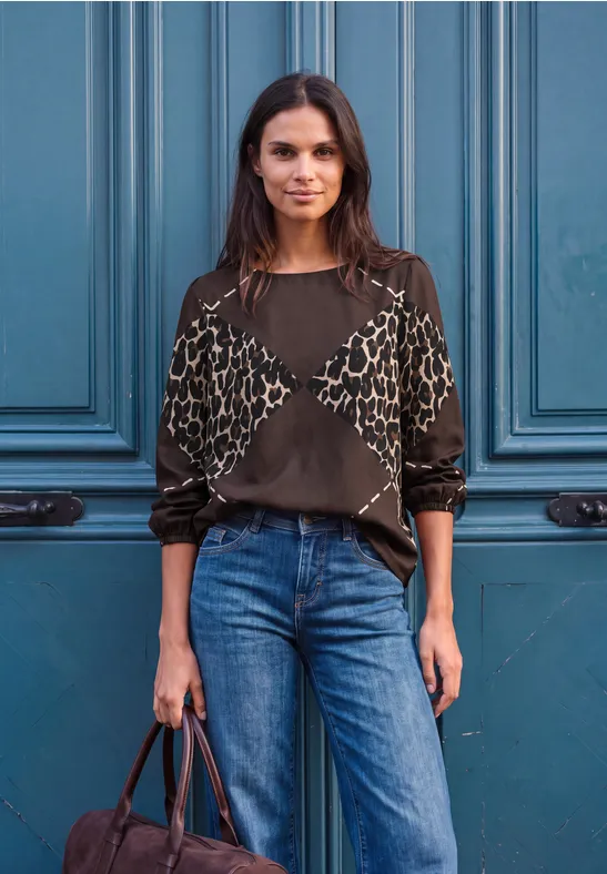 Eine Frau steht selbstbewusst vor einer blauen Tür, trägt Jeans und eine gemusterte Bluse.
