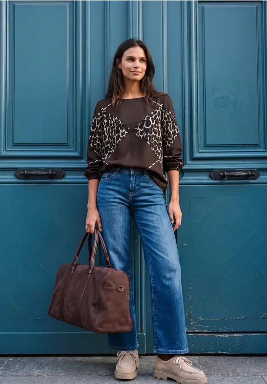 Eine Frau steht vor einer blauen Tür, trägt weite Jeans, gemusterten Pullover und hält eine braune Tasche.