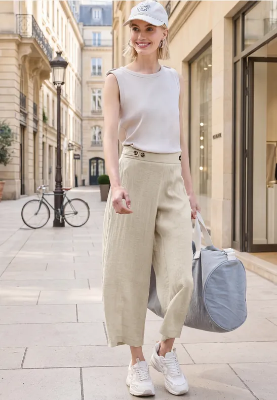 Frau in weißem Top, weiten beigen Hosen und Sneakers mit Reisegepäck schlendert durch eine elegante Straßenkulisse.
