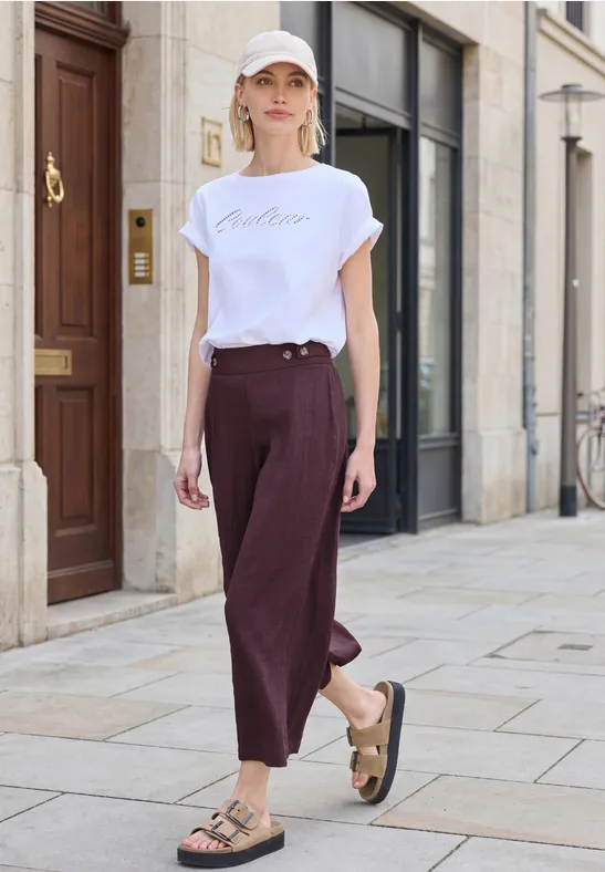 Person in weißem T-Shirt und weinroter Hose mit Sandalen spaziert auf einem Gehweg.