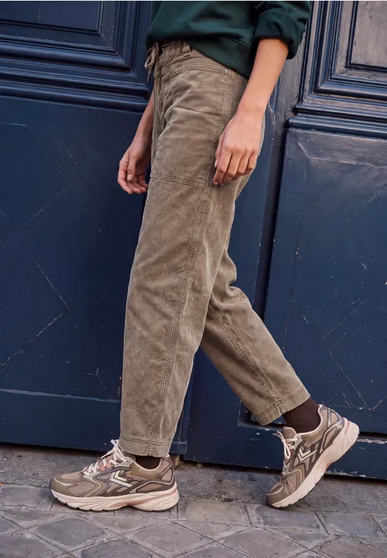 Eine Person trägt beige Cordhosen und braune Sneaker vor einer dunklen Tür