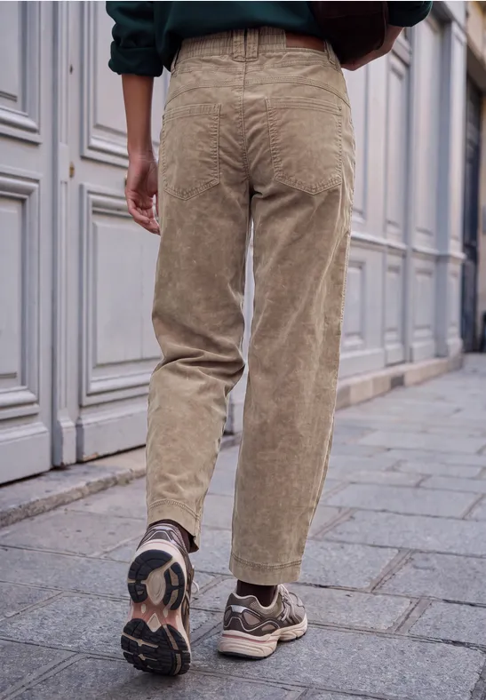Rückansicht einer Person in beigen Cordhose und Turnschuhen auf gepflastertem Gehweg vor einer Hausfassade.