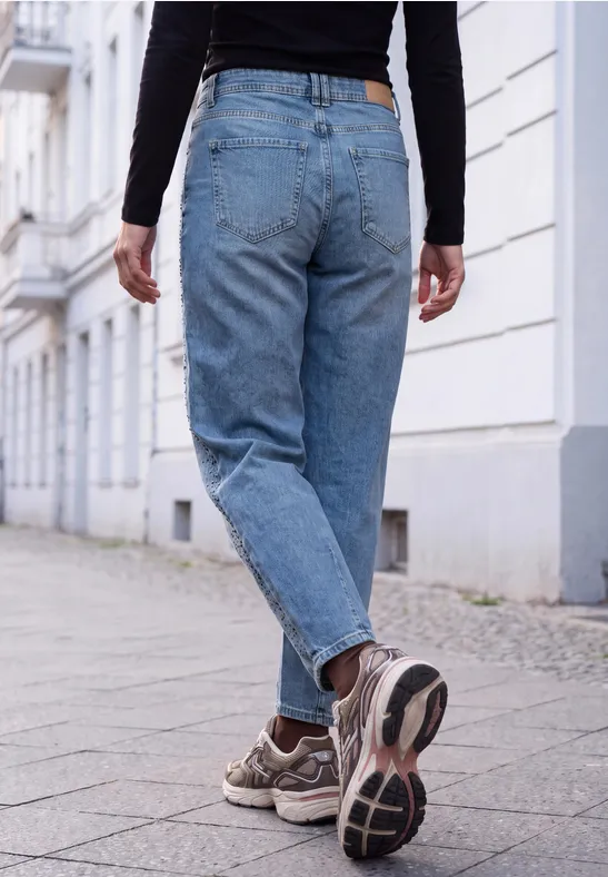 Widok od tyłu osoby w niebieskich dżinsach i sneakersach, idącej chodnikiem w cichej, miejskiej okolicy.