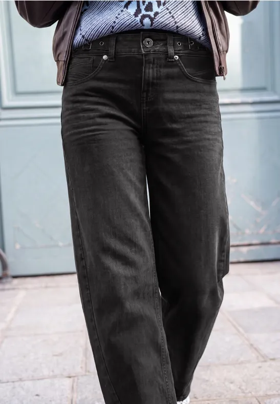 Schwarze, gerade geschnittene Jeans an einer Person, kombiniert mit gemustertem Hemd vor blauer Tür.