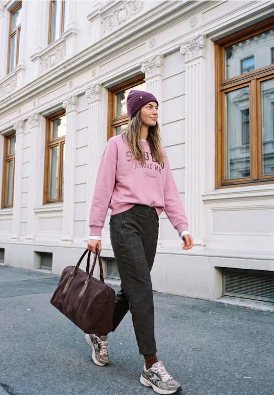Junge Frau in pinkem Hoodie und Mütze spaziert mit Tasche vor klassischer Fassade.