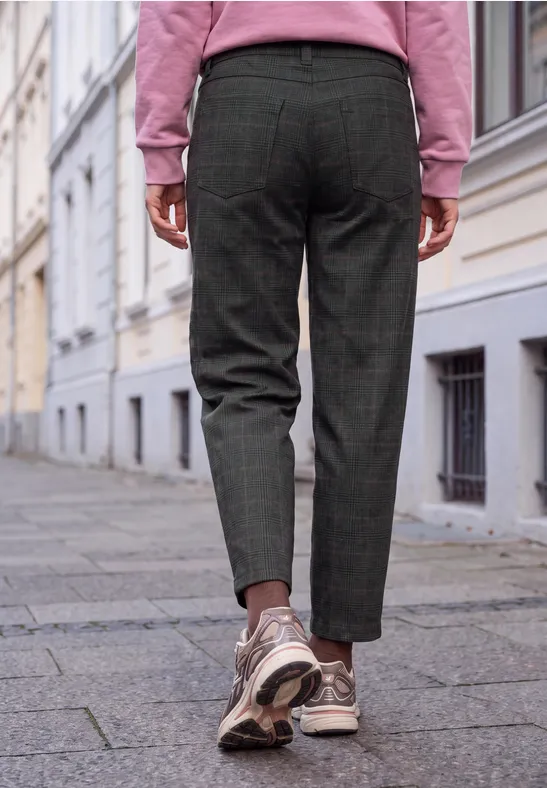Eine Person in karierter Hose, rosa Pulli und Sneakers geht auf einem Gehweg an Häusern vorbei.