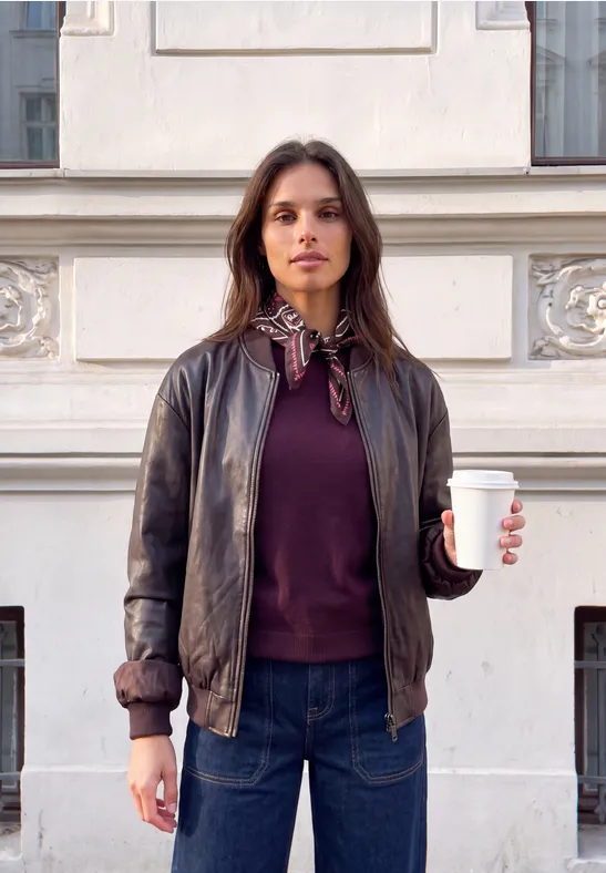 Frau trägt Lederjacke und Jeans, steht vor einem Gebäude, hält einen Kaffee zum Mitnehmen.