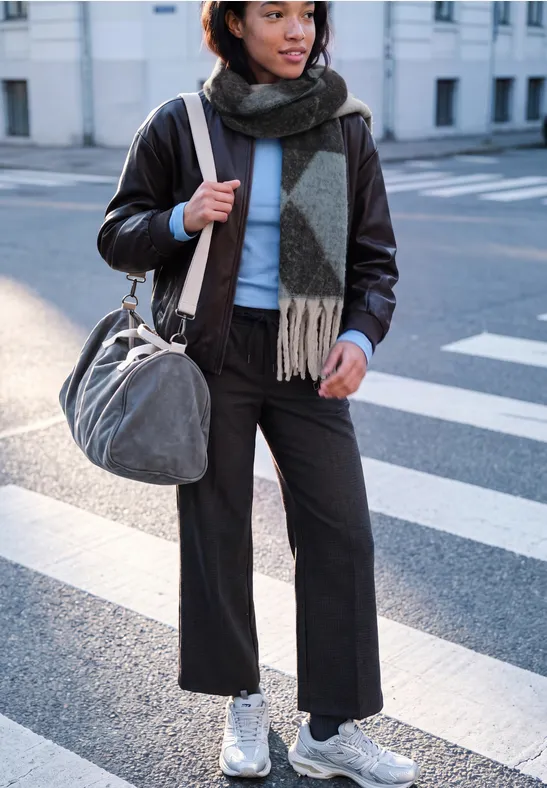 Frau in Lederjacke mit Schal und Tasche überquert Straße in weißen Sneakers.