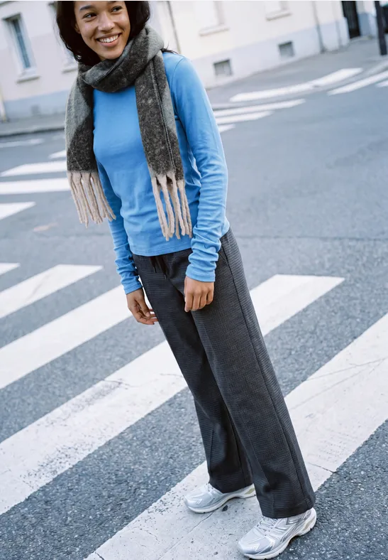 Junge Frau lächelt auf Zebrastreifen, trägt blauen Pullover, grauen Schal, weite Hose und weiße Sneaker.