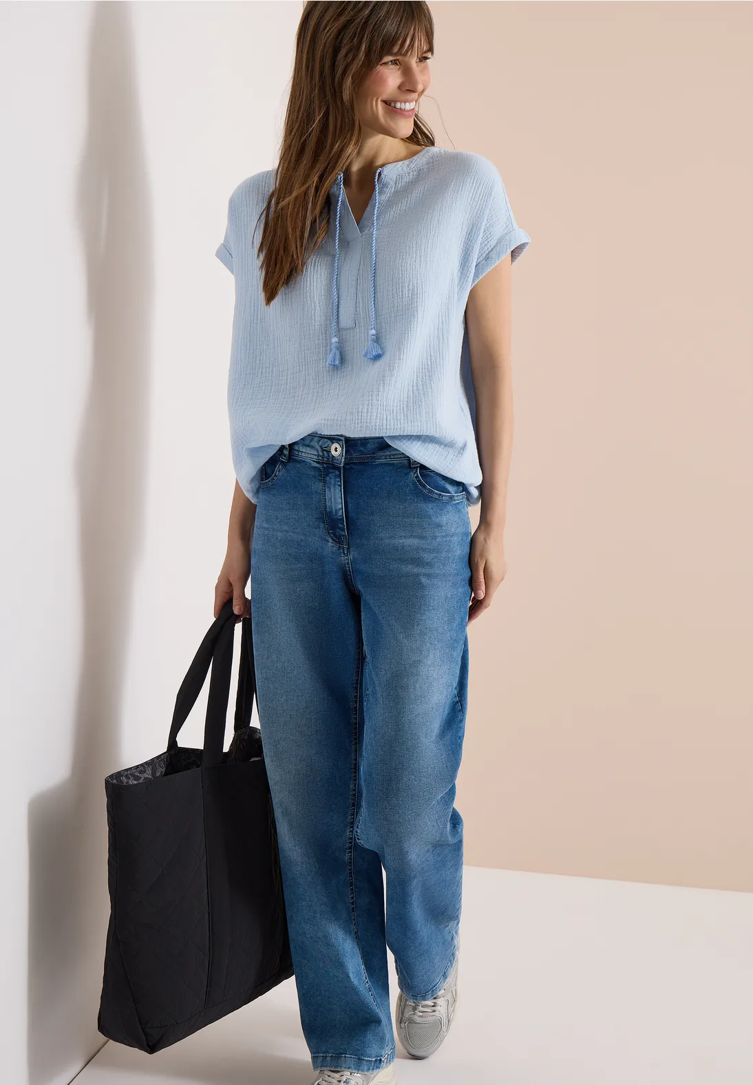 Eine Frau trägt hellblaues T-Shirt, weite blaue Jeans, weiße Sneaker und eine schwarze Tragetasche.