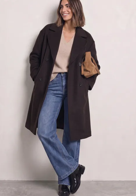 Frau in dunklem Mantel, beigem Pullover, weiten Jeans und schwarzen Stiefeln hält eine Clutch.