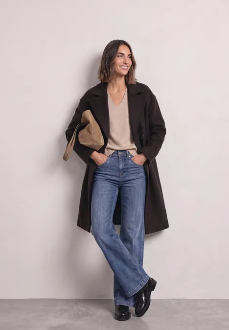 Frau in braunem Mantel, beigem Top, weiten Jeans und schwarzen Schuhen vor hellem Hintergrund.