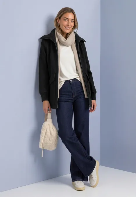 Mantel mit Wollanteil und Strickkragen Black Frau in weißem Pullover, Schal und dunkler Jacke, mit Schlagjeans, weißen Sneakern und heller Tasche vor blauer Wand.