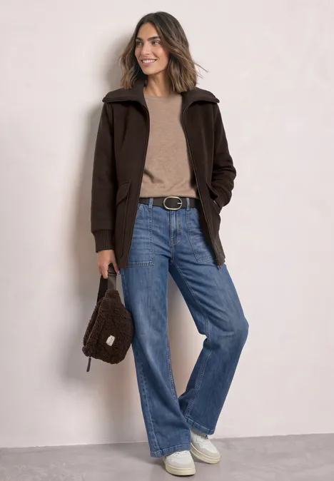 Mantel mit Wollanteil und Strickkragen auburn brown Frau in brauner Jacke, beigem Pullover, blauen Jeans und weißen Sneakern, hält eine braune Tasche.