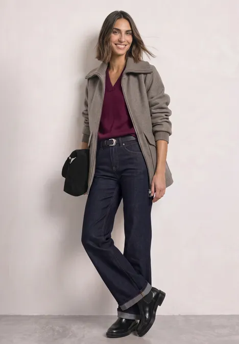 Frau in grauer Jacke, Bordeaux-Shirt und weiten Jeans, schwarze Stiefel und Tasche, lächelnd stehend.