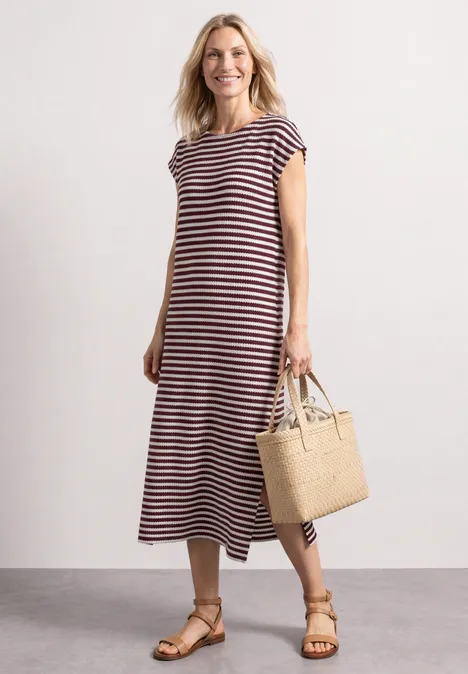 Frau trägt gestreiftes Midikleid, Sandalen und Basttasche vor schlichtem Hintergrund.