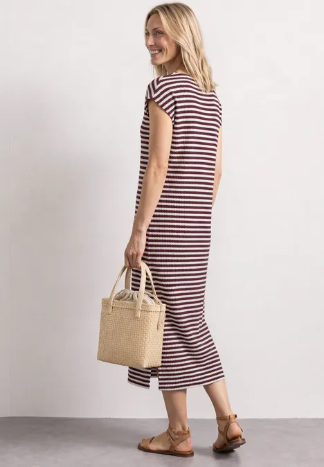 Frau in rot-weiß gestreiftem Midikleid mit Strohtasche und Sandalen vor hellem Hintergrund.