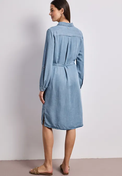 Vrouw van achteren in een knielange spijkerjurk met lange mouwen, blootsvoets.