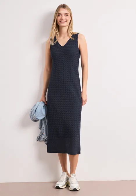 Vrouw in mouwloze donkerblauwe midi-jurk met ruitpatroon, witte sneakers en spijkerjack los over de schouder.