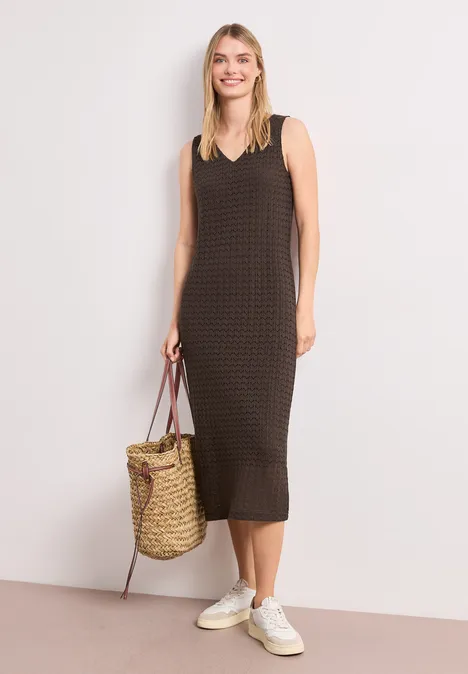 Vrouw in mouwloze midi-jurk met fijn patroon, witte sneakers en een strooien tas.