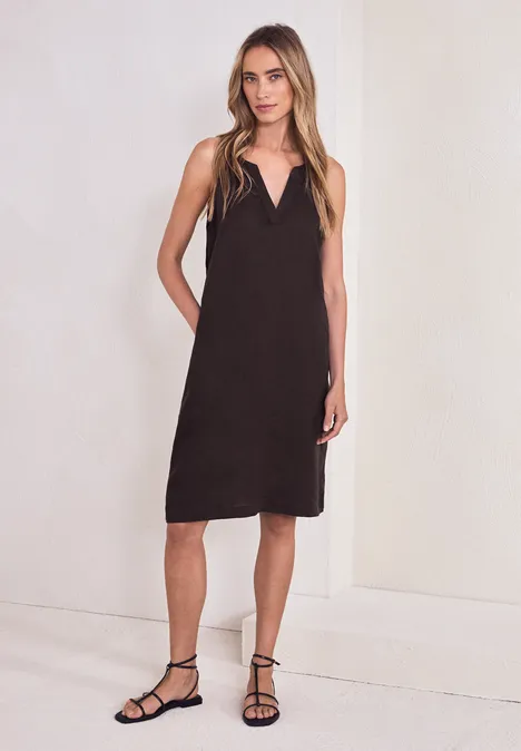 Femme en robe mi-longue noire sans manches et sandales fines noires devant un fond clair.