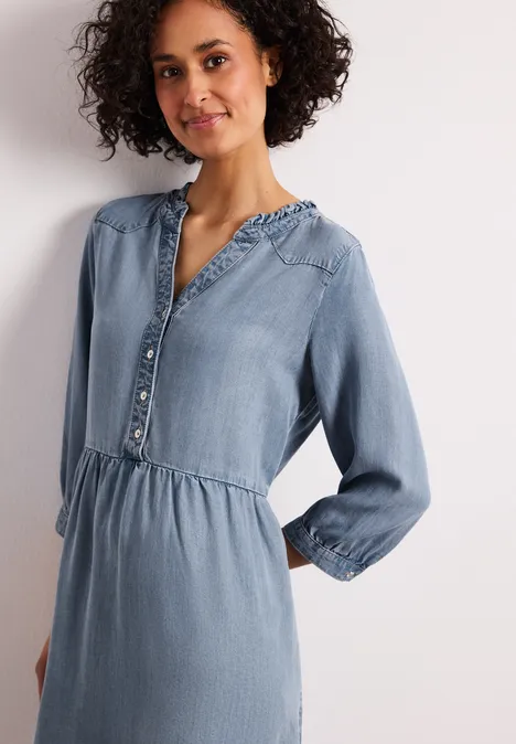 Frau in hellblauem Jeanskleid mit Knopfleiste, elastischer Taillennaht und langen Ärmeln.