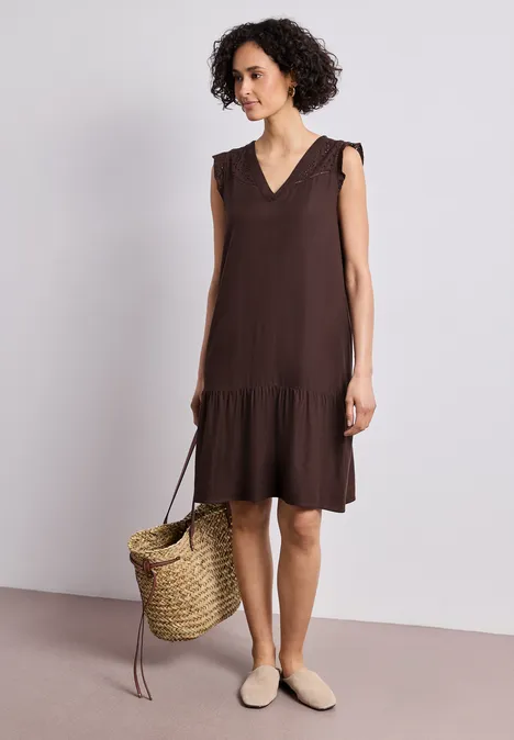 Frau mit braunem ärmellosem Kleid mit Stufensaum, Strohtasche und hellen Slippern vor neutralem Hintergrund.