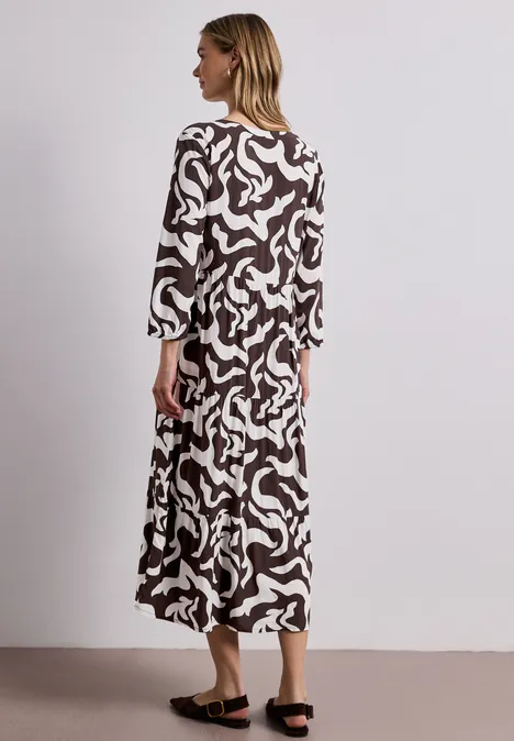 Frau im langen, fließenden Kleid mit braun-weißem abstraktem Muster vor neutralem Hintergrund.
