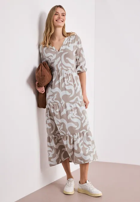 Eine Frau in fließendem, gemustertem Midikleid lehnt an einer weißen Wand, dazu weiße Sneaker und Clutch.