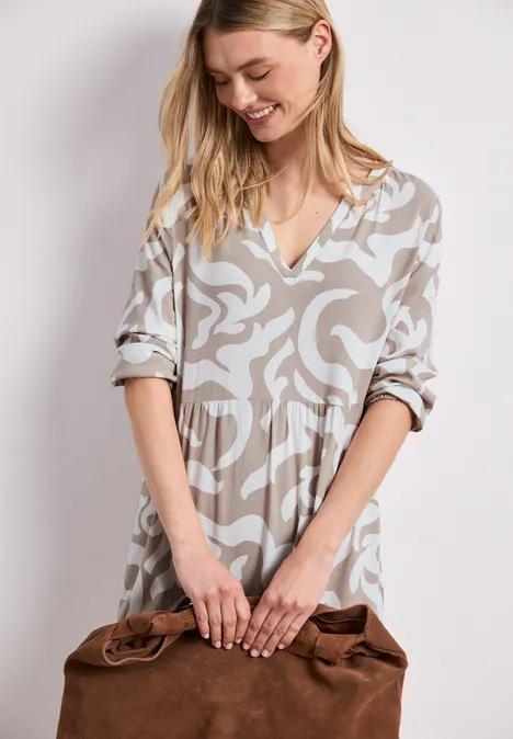 Frau in beige-gemustertem Midikleid mit V-Ausschnitt und 3/4-Ärmeln hält eine braune Ledertasche.