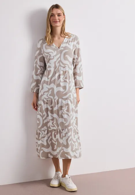Frau trägt ein beiges Midi-Kleid mit weißem, abstraktem Muster, V-Ausschnitt und weißen Sneakern