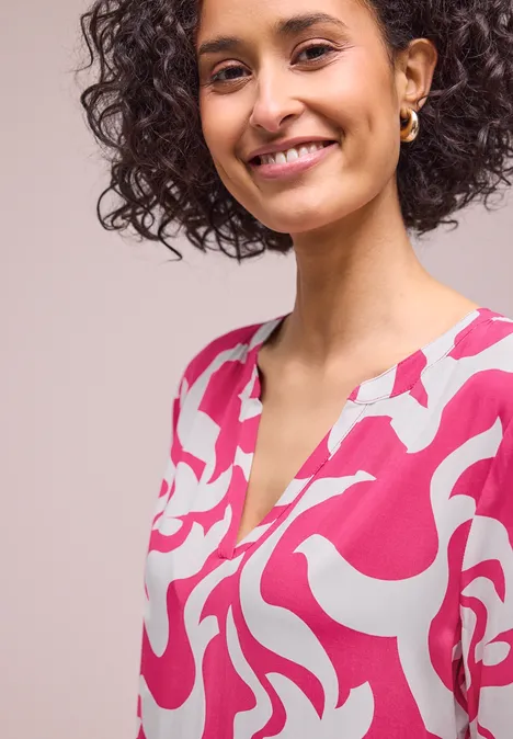 Eine Person trägt eine pink-weiße, abstrakt gemusterte Bluse, mit lockigem Haar und goldener Ohrring.
