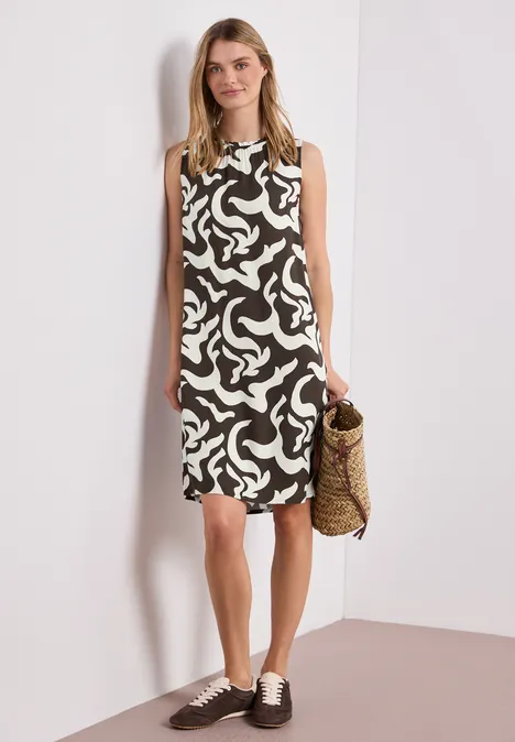 Frau trägt ärmelloses, schwarz-weiß gemustertes Kleid, hält Strohkorb, trägt braune Schnürschuhe