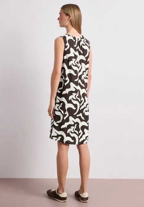 Rückenansicht einer Frau im ärmellosen, schwarz-weißen Kleid mit abstraktem Muster und flachen Slip-on-Schuhen.