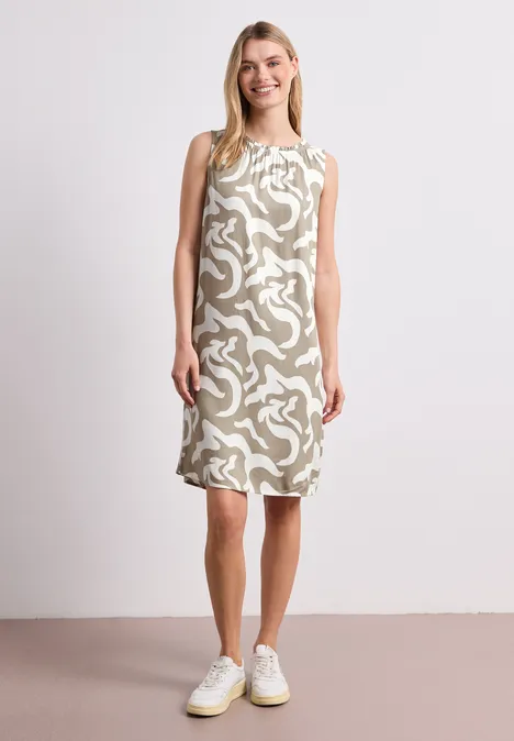 Frau in ärmellosem, knieumspielendem Kleid mit abstraktem Muster trägt weiße Sneaker vor neutralem Hintergrund.