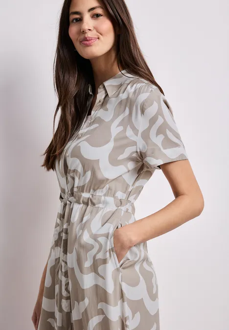 Femme de profil porte une robe-chemise gris clair à motif blanc, manches courtes, taille ajustée et poches.