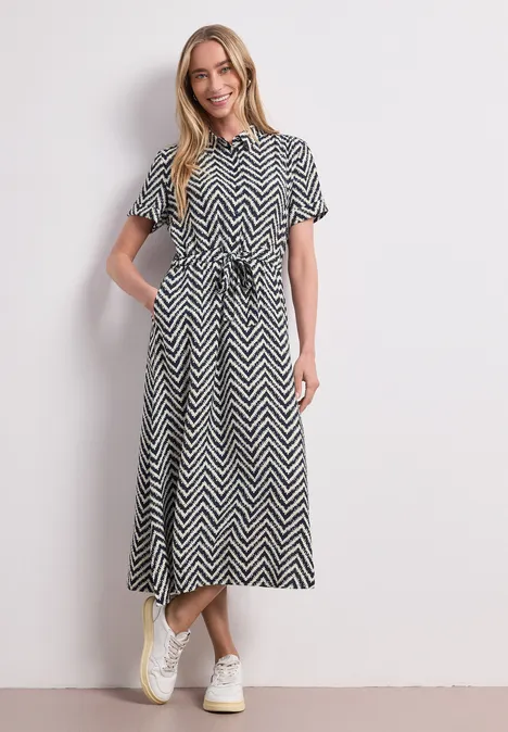 Femme en robe midi à chevrons noir et blanc avec ceinture, baskets blanches et une main dans la poche.