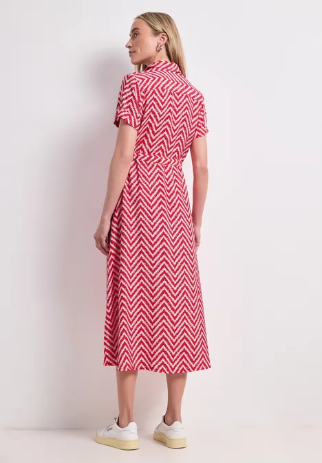 Femme de profil portant une robe mi-longue rouge à motifs chevrons blancs et baskets blanches sur fond clair.