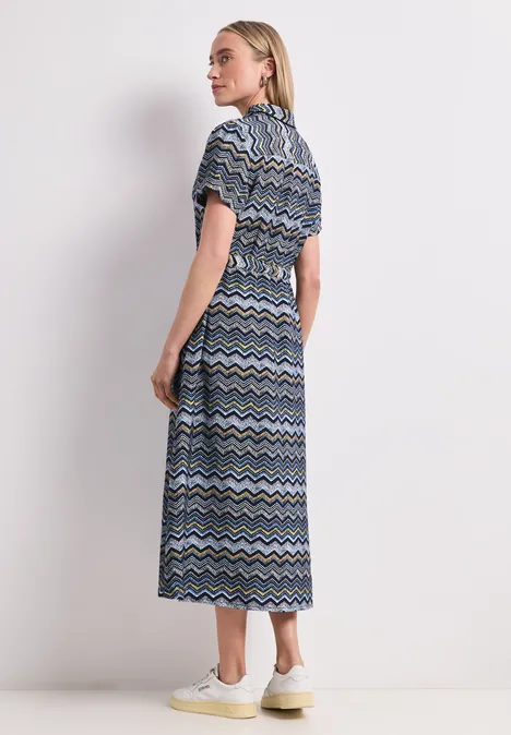 Frau in midi Kleid mit blauem, weißem und schwarzem Zickzackmuster steht seitlich vor weißem Hintergrund.