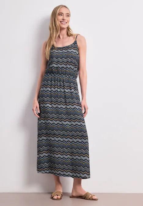 Frau in langem, ärmellosem Kleid mit buntem Zickzackmuster, Sandalen und weißem Hintergrund.