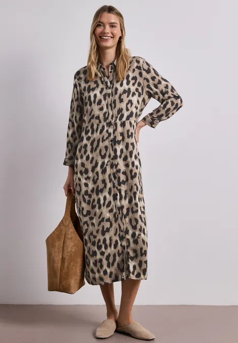 Vrouw in lange luipaardjurk met grote bruine tas en beige schoenen.
