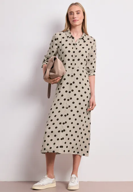 Vrouw in beige midijurk met zwart hartpatroon, witte sneakers en beige schoudertas.