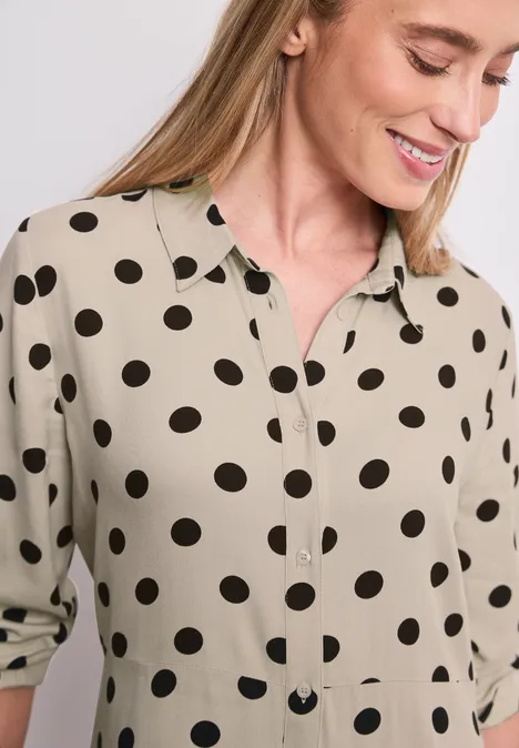 Beige blouse met zwarte polkadots, kraag en knoopsluiting, mouwen nonchalant opgerold.