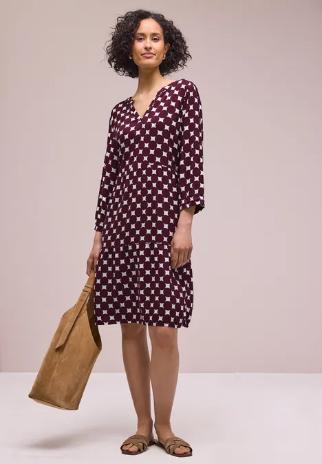 Femme en robe mi-longue bordeaux à pois blancs, manches 3/4, portant un sac marron et des sandales plates.