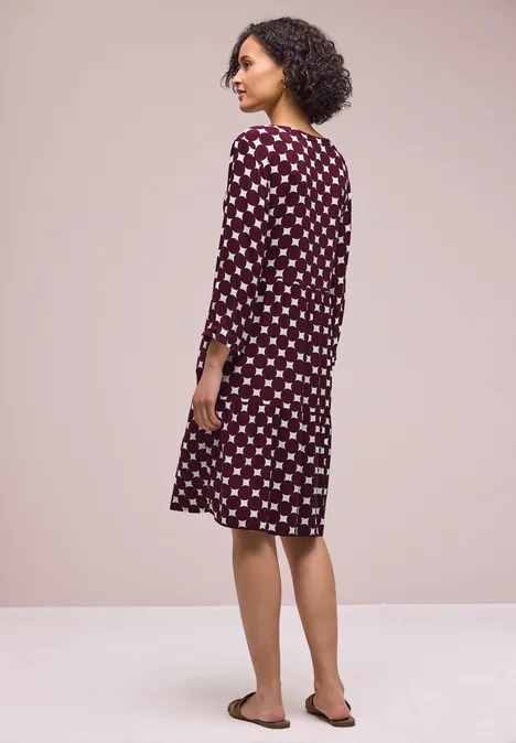 Une personne aux cheveux bouclés vue de dos porte une robe bordeaux à pois blancs et des sandales devant un mur beige.